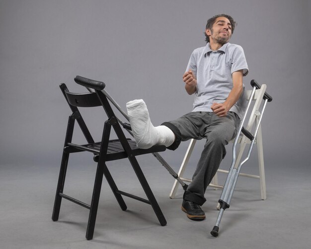 正面図灰色の壁に壊れた足と松葉杖で座っている若い男性壊れた事故足足の痛みのねじれ