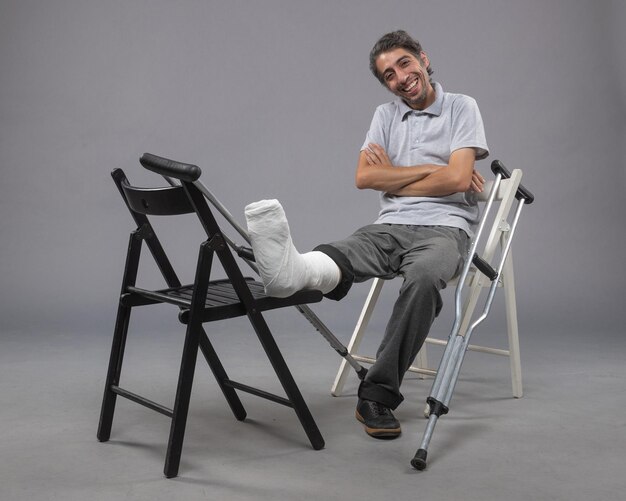 Front view young male sitting with broken foot and crutches smiling on grey wall accident twist leg pain broken foot