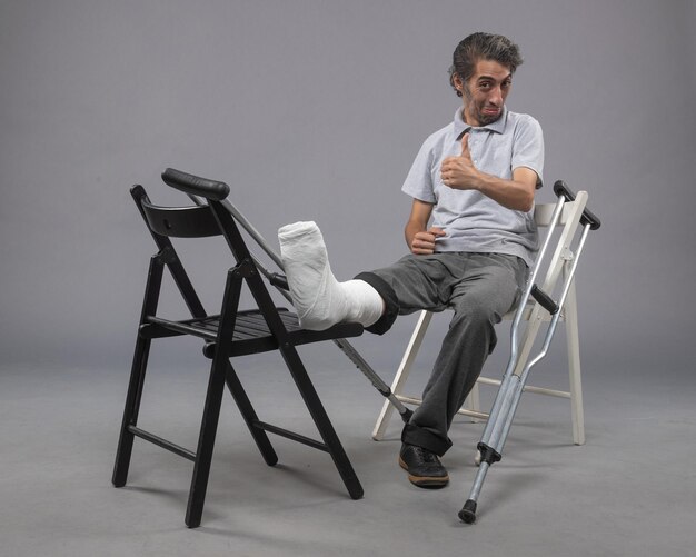 正面図若い男性が壊れた足と松葉杖で灰色の壁に笑みを浮かべて座っている事故足のねじれの痛み足の骨折