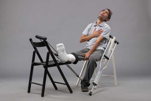 Vista frontale giovane maschio seduto con le stampelle del piede rotto e che soffre di dolore sul muro grigio dolore al piede incidente gamba rotta torsione