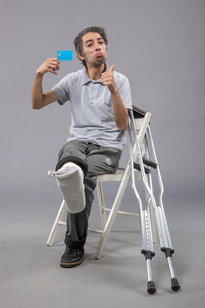 Front view young male sitting with broken foot holding blue bank card on grey floor pain foot broken accident twist male