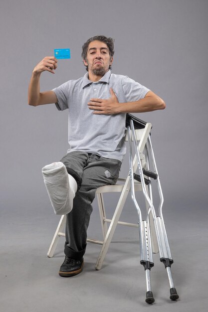 Front view young male sitting with broken foot holding blue bank card on grey wall broken accident foot pain twist male leg