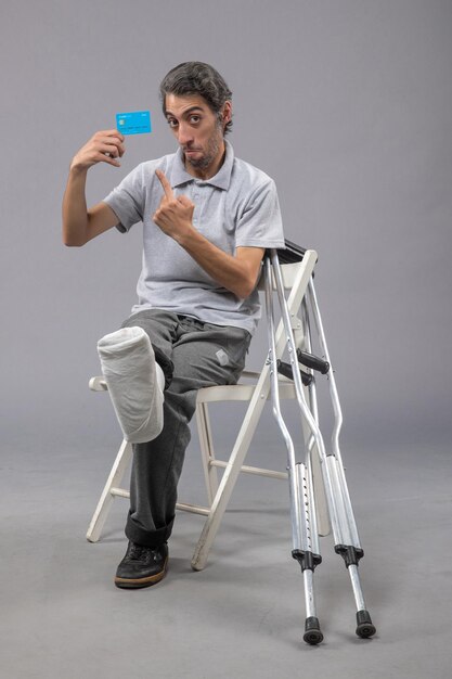 Front view young male sitting with broken foot holding blue bank card on grey wall male pain foot broken crutch accident leg