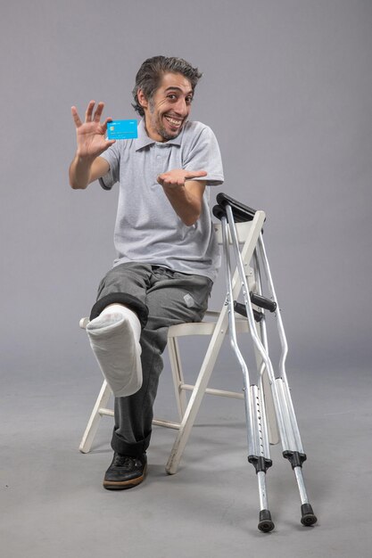 灰色の壁に青い銀行カードを保持している壊れた足で座っている正面図若い男性痛み足壊れた事故脚のねじれ