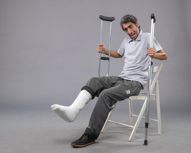 Front view young male sitting with broken foot and holding crutches on grey wall foot broken accident leg pain twist