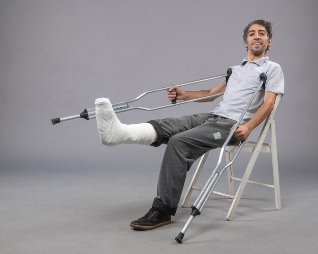 Front view young male sitting with broken foot and holding crutches on grey wall pain foot broken accident leg twist