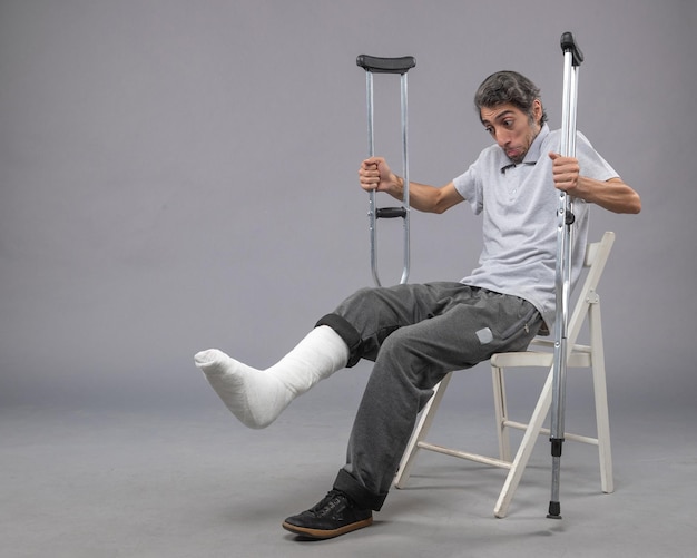 Free photo front view young male sitting with broken foot and holding crutches on grey wall pain foot broken accident legs twist