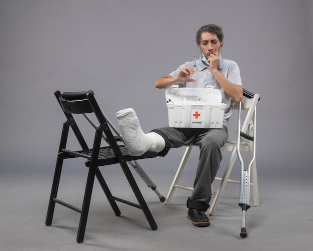 Free photo front view young male sitting with broken foot and holding injection on a grey wall medicine twist male pain foot leg broken