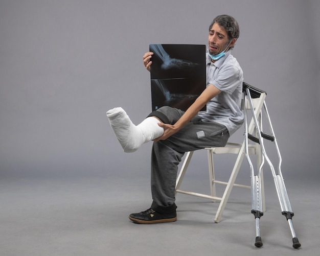 Free photo front view young male sitting with broken foot and holding x-ray of it on grey desk pain leg broken male accident foot twist