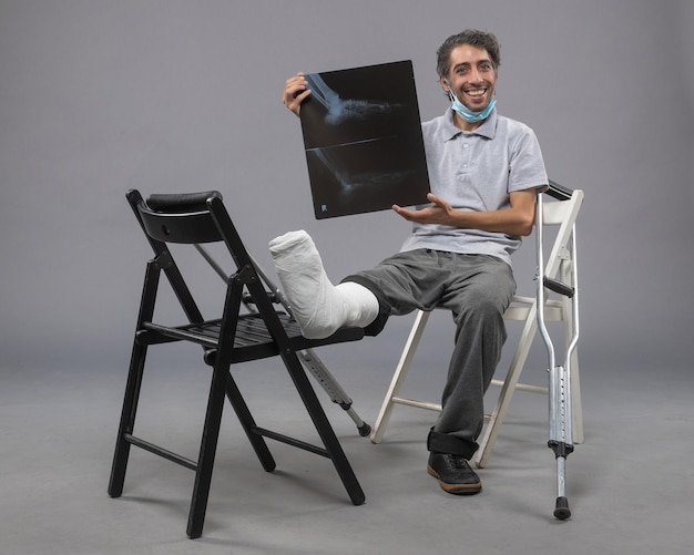 Foto gratuita vista frontale giovane maschio seduto con il piede rotto e tenendo i raggi x su pavimento grigio dolore alla torsione della gamba del piede rotto incidente maschile