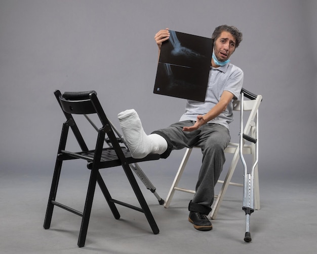 Free photo front view young male sitting with broken foot and holding x-ray of it on grey floor twist pain leg broken male accident foot