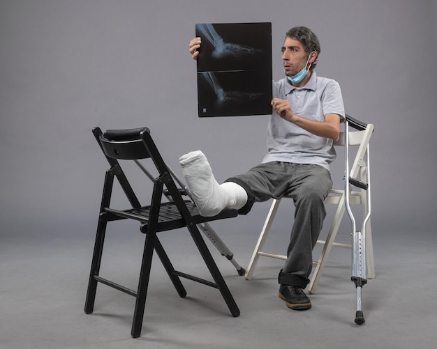 Вид спереди молодого мужчины, сидящего со сломанной ногой и держащего рентгеновский снимок на серой стене.