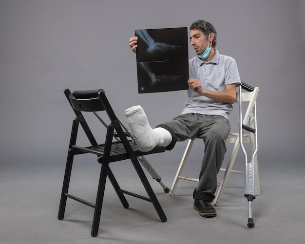 Вид спереди молодого мужчины, сидящего со сломанной ногой и держащего рентгеновский снимок на серой стене.