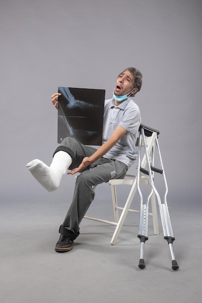 Front view young male sitting with broken foot and holding x-ray of it on a grey wall pain leg accident foot twist male