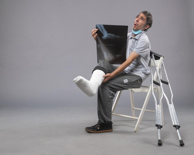 Front view young male sitting with broken foot and holding x-ray of it on a grey wall pain leg broken accident foot twist male