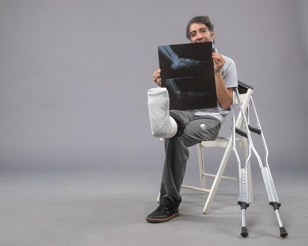 Вид спереди молодого мужчины, сидящего со сломанной ногой и держащего рентгеновский снимок на серой стене, боль в ноге, сломанная мужская авария, скручивание стопы
