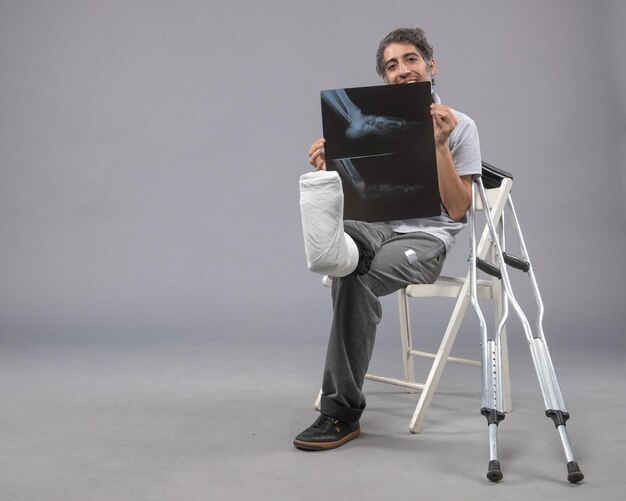 Front view young male sitting with broken foot and holding x-ray of it on grey wall pain leg broken male accident foot twist