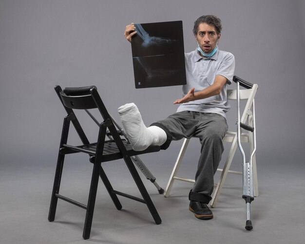 Front view young male sitting with broken foot and holding x-ray of it on grey wall twist pain foot leg broken male accident