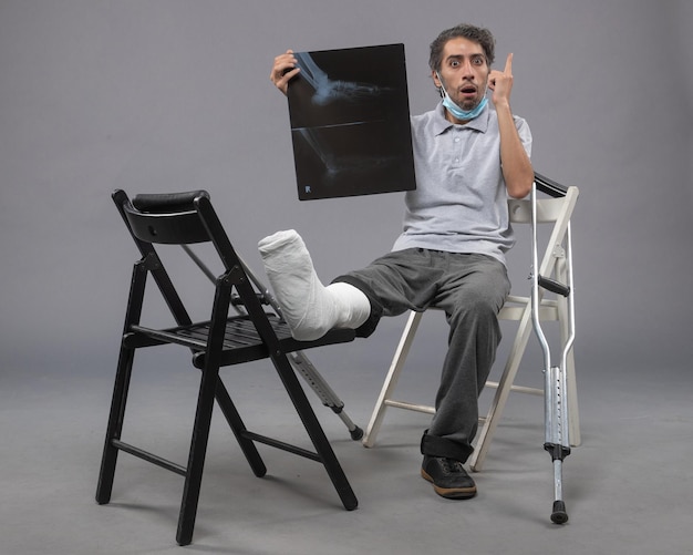 Free photo front view young male sitting with broken foot and holding x-ray of it on the grey wall twist pain foot leg broken male accident