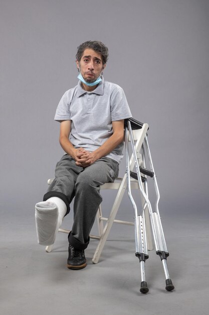 Front view young male sitting with broken foot and tied bandage on the grey wall foot twist male accident pain