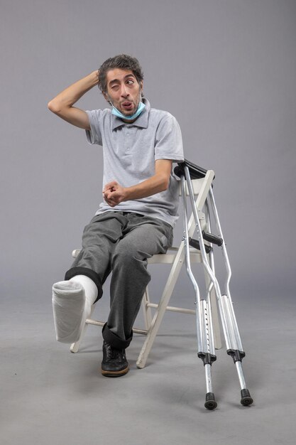 Front view young male sitting with broken foot and tied bandage on grey wall pain foot twist male accident