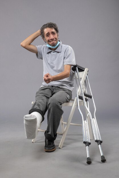 Front view young male sitting with broken foot and tied bandage on the grey wall pain foot twist male accident