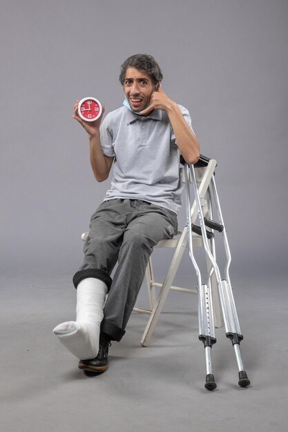 Front view young male sitting with broken foot and tied bandage holding clock on a grey wall foot twist male accident pain leg