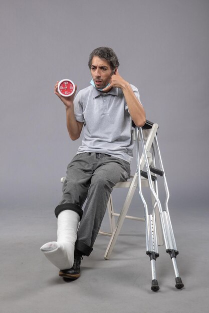 Front view young male sitting with broken foot and tied bandage holding clock on grey wall foot twist male accident pain leg