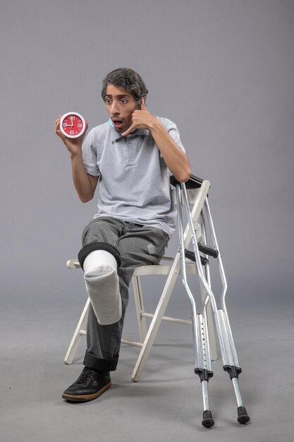 Front view young male sitting with broken foot tied bandage and holding clock on grey wall foot twist male accident pain leg