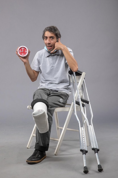 Front view young male sitting with broken foot tied bandage and holding clock on grey wall foot twist male accident pain legs