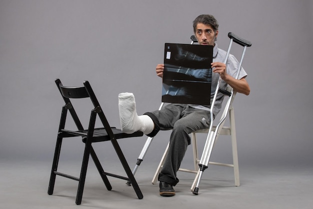 Бесплатное фото Вид спереди молодого мужчины, сидящего со сломанной ногой, используя костыли и держащего рентгеновский снимок на серой стене, скручивание стопы, сломанная боль, авария