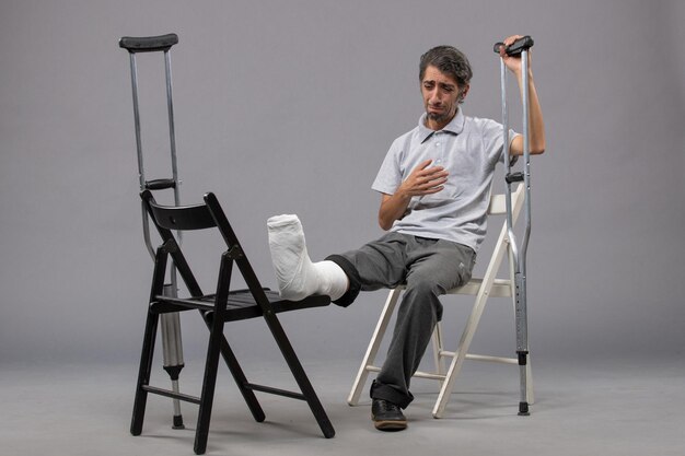 Front view young male sitting with broken foot and using crutches feeling sad on grey wall twist broken pain foot accident leg