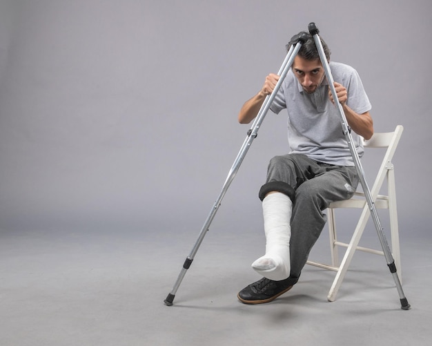 Бесплатное фото Вид спереди молодого мужчины, сидящего со сломанной ногой и использующего костыли для ходьбы на сером столе, скручивание ноги, авария, боль, сломанная стопа