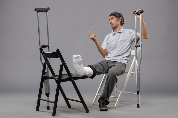 Front view young male sitting with broken foot and using crutches on grey desk twist broken pain foot accident leg