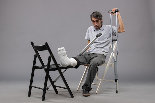 正面図若い男性が足を骨折して座って、灰色の床に松葉杖を使用して足をひねる痛み事故の足を骨折