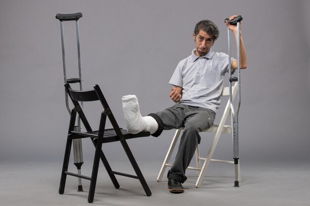 Front view young male sitting with broken foot and using crutches on grey wall foot twist broken pain accident legs