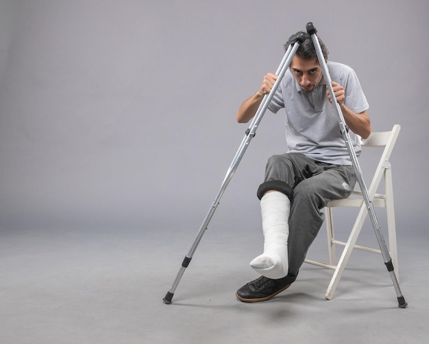 Vista frontale giovane maschio seduto con il piede rotto usando le stampelle per camminare sulla scrivania grigia gamba torsione incidente dolore piede rotto