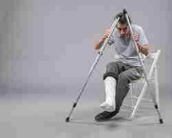 Free photo front view young male sitting with broken foot using crutches for walking on grey desk leg twist accident pain broken foot