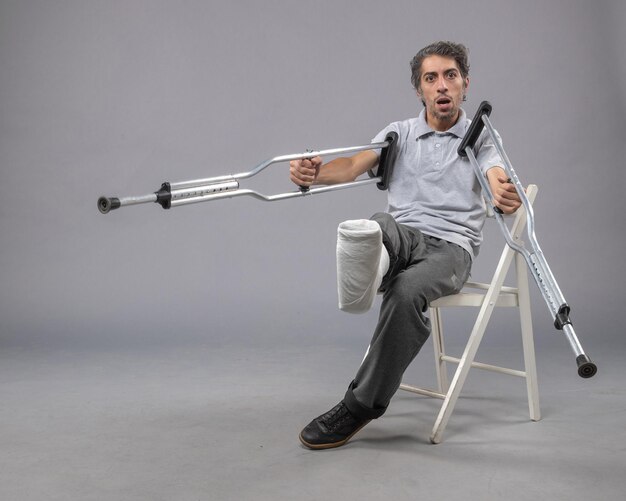 Front view young male sitting with broken foot using crutches for walking on grey wall foot twist accident legs pain broken