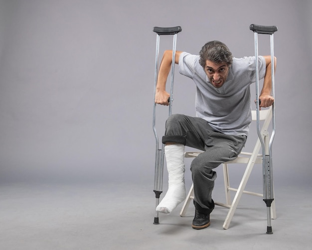 Free photo front view young male sitting with broken foot using crutches for walking on grey wall leg twist accident pain broken