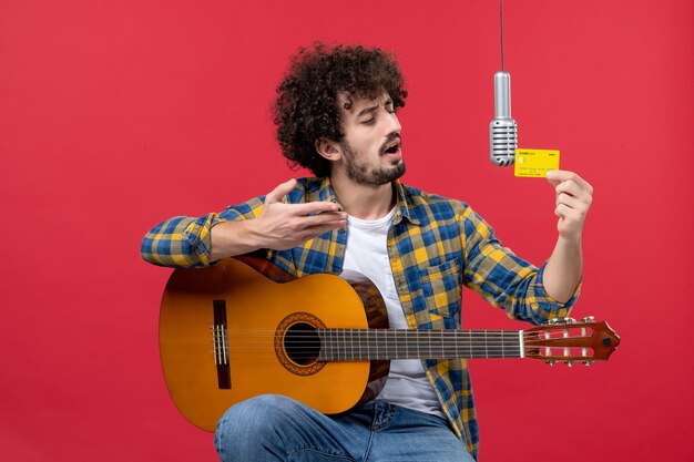 Вид спереди молодой мужчина, сидящий с гитарой, держащий банковскую карту на красной стене, аплодисменты, музыкант, играющая группа, концертная музыка, живая