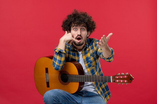 Front view young male sitting with guitar on red wall applause band concert musician play music live