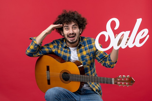 Giovane maschio di vista frontale che si siede con la chitarra sul musicista di applauso di colore della parete rossa che suona la vendita di musica di concerto della banda