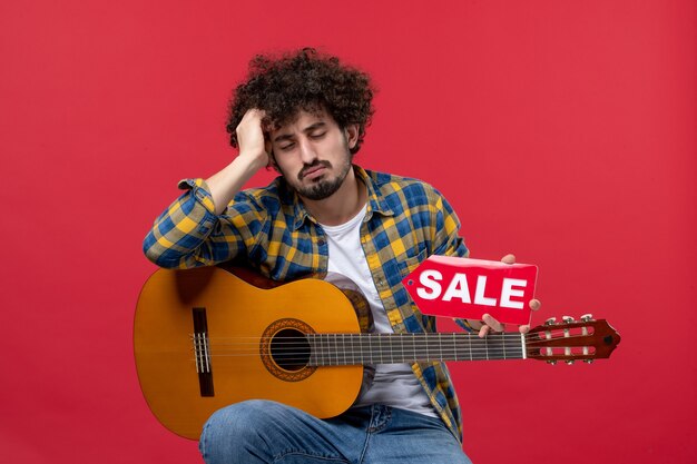 Вид спереди молодой мужчина, сидящий с гитарой на красной стене, аплодисменты, музыкант, играющий группу, музыка, живая продажа