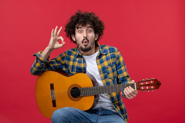 Front view young male sitting with guitar on red wall concert live color band music play musician applause