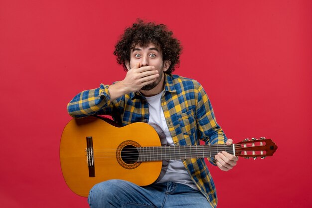 Front view young male sitting with guitar on red wall live applause musician play color band concert music