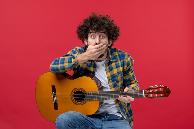 Foto gratuita vista frontale giovane maschio seduto con la chitarra sul muro rosso suona musica da concerto musicista colore applausi live band