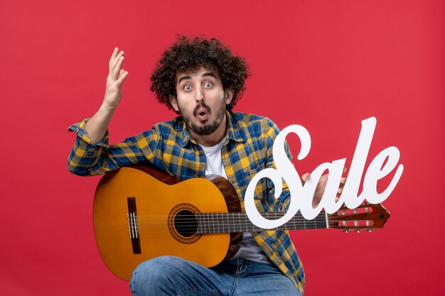 Вид спереди молодой мужчина, сидящий с гитарой на красной стене, играет на концерте, продажа музыкантов, цветная музыка, аплодисменты вживую
