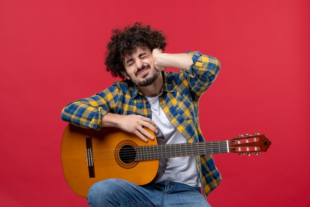 Front view young male sitting with guitar on red wall play live music color musician applause band