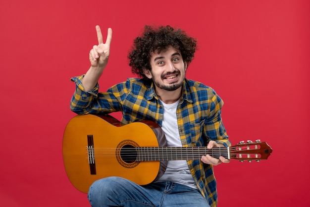 Front view young male sitting with guitar on red wall play music musician color applause live concert